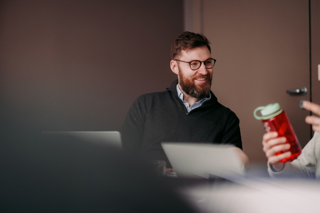 A person wearing glasses and a dark sweater sits at a table with a laptop, smiling at another individual holding a red water bottle.
