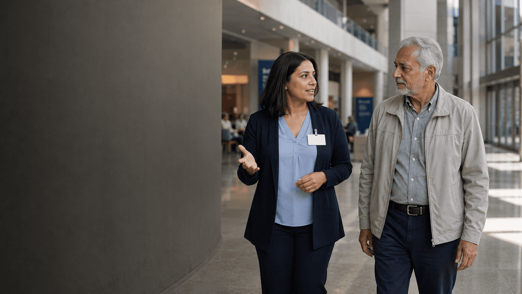 A hospital coordinator helps an older visitor find his way through a bright hospital atrium.