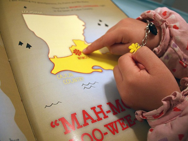 A child's hands, wearing a pink long-sleeve shirt with small hearts, hold a small yellow flower and a chain with a small flower charm. The child points to a yellow shape on the page of a book. The page shows a map of Louisiana and the words “CAJUN COUNTRY” are printed below it. A red quotation mark appears below the title “MAH-MOO-WEE”. The map includes small airplane icons and stylized waves.