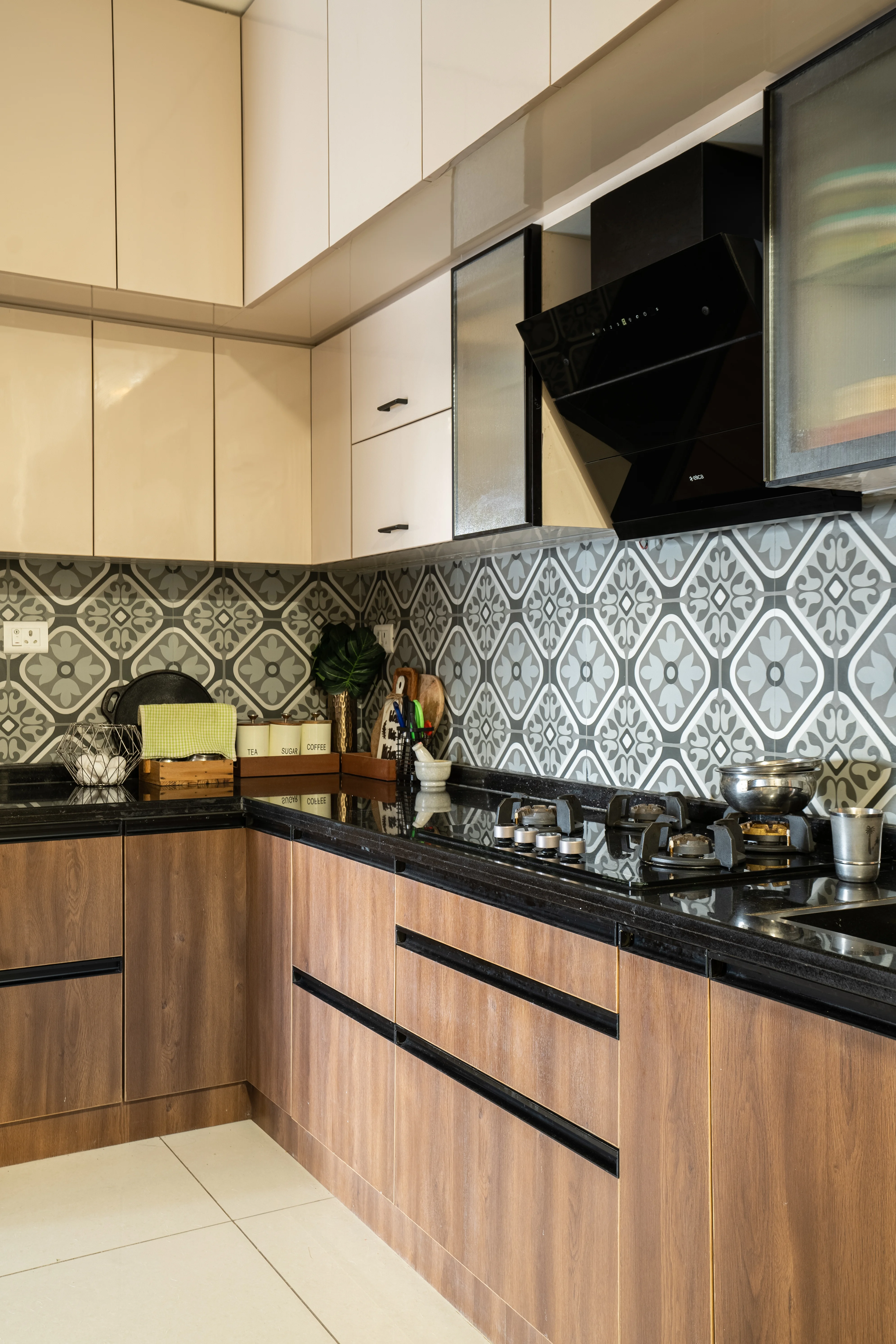 A modern kitchen with wooden cabinets, a black countertop, and morrocan tile backsplash. A black range hood hangs above the stove, and there are various cooking utensils and appliances on the countertop.