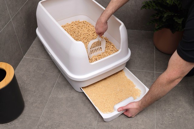 A person cleans a white, Treasured Pals Premium Sifting Litter System with a scoop, removing waste to reveal fresh litter pellets, on a gray tiled floor next to a potted plant.
