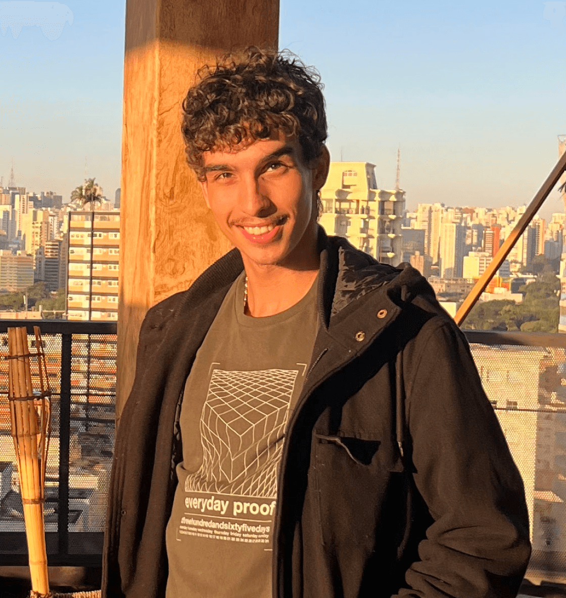 A man with short curly hair wearing a jacket, smiling under a sunny sky with buildings visible behind him.