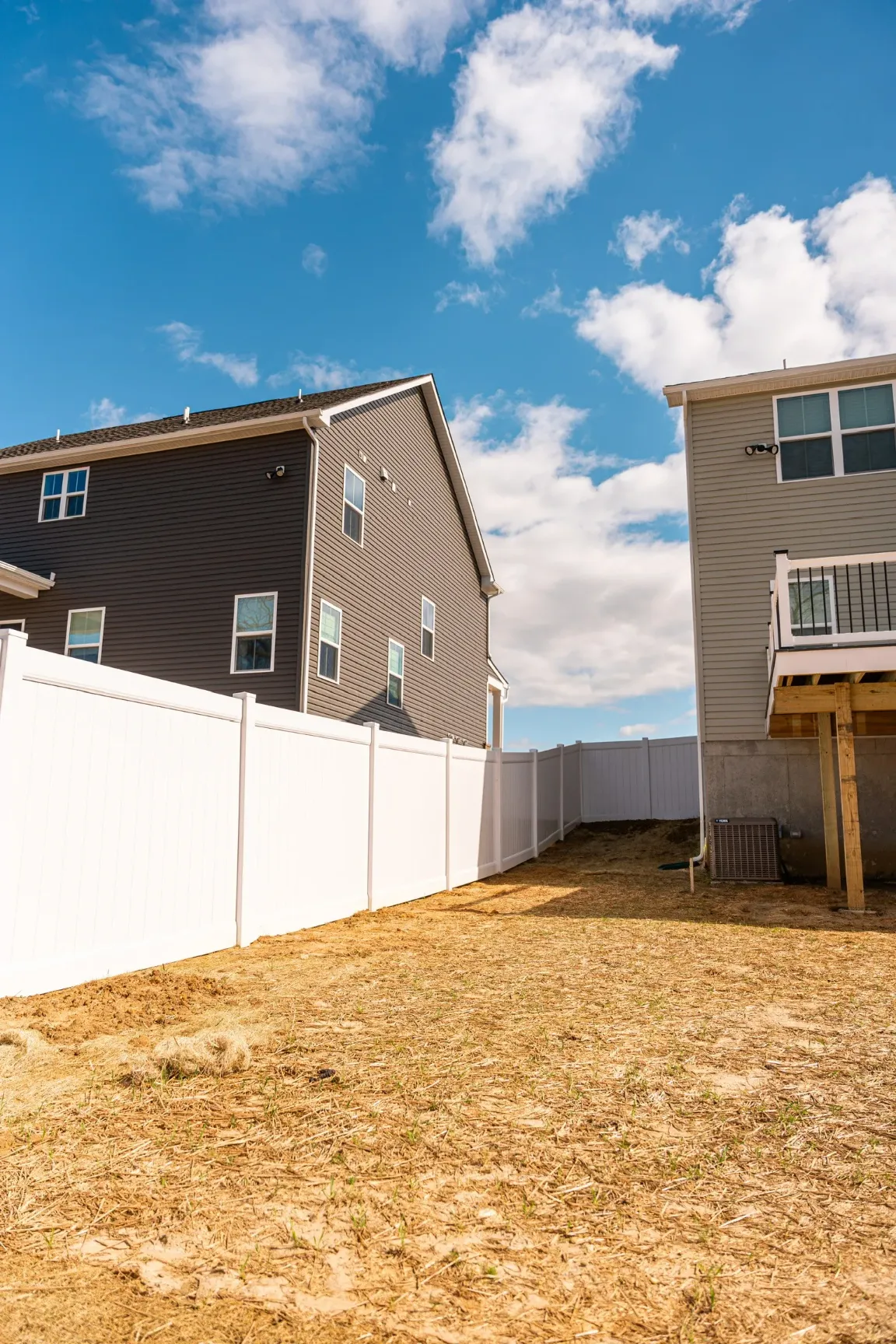 A close-up of a modern outdoor railing post with clean lines and a soft, warm LED light trim, set against the backdrop of a house featuring large windows and a black grill on a patio.