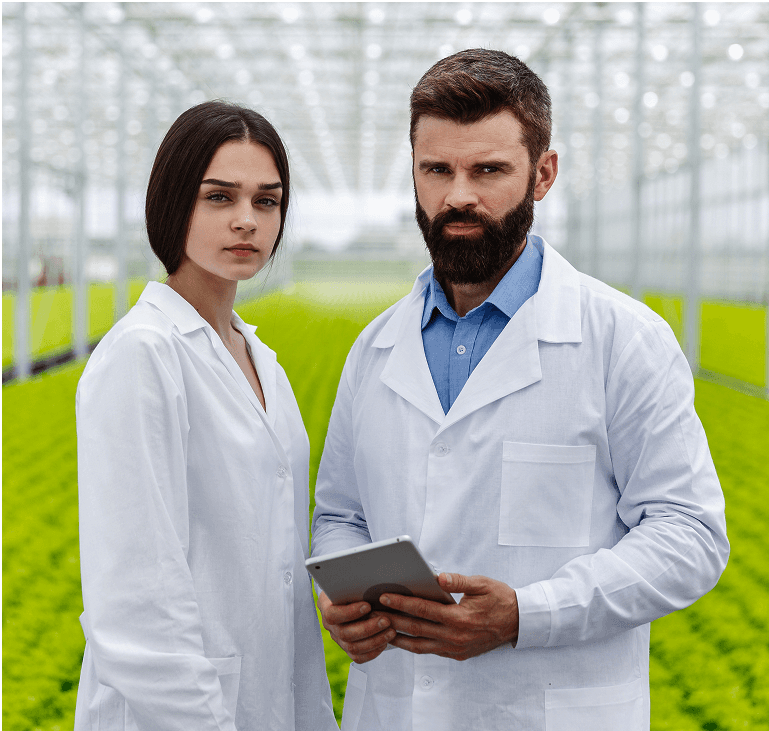 Two scientists in white lab coats stand in a greenhouse filled with lush green plants, with one holding a tablet, demonstrating agricultural research and technology.