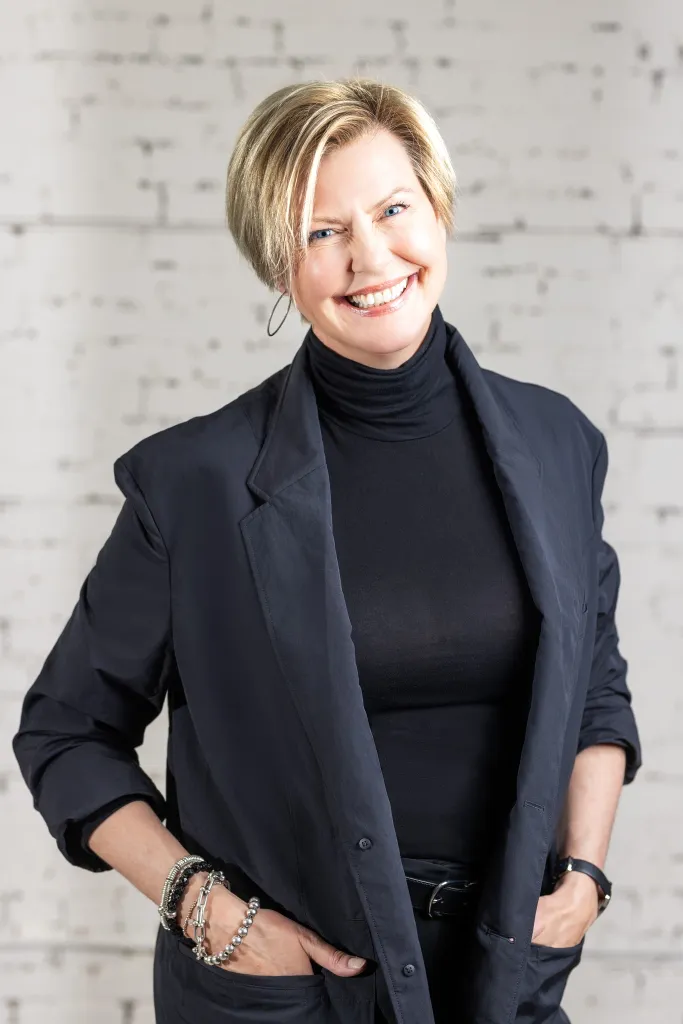 Image of Blonde woman smiling, dressed in black, standing against a white brick wall background, representing KHD Homes.