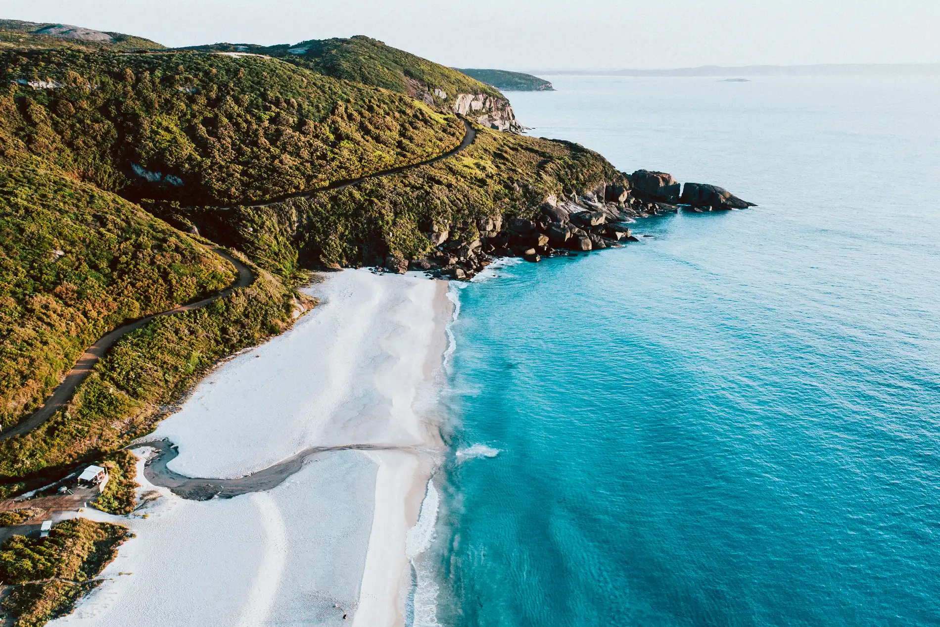 Aerial of a green headland dropping to a white sand beach and deep blue water