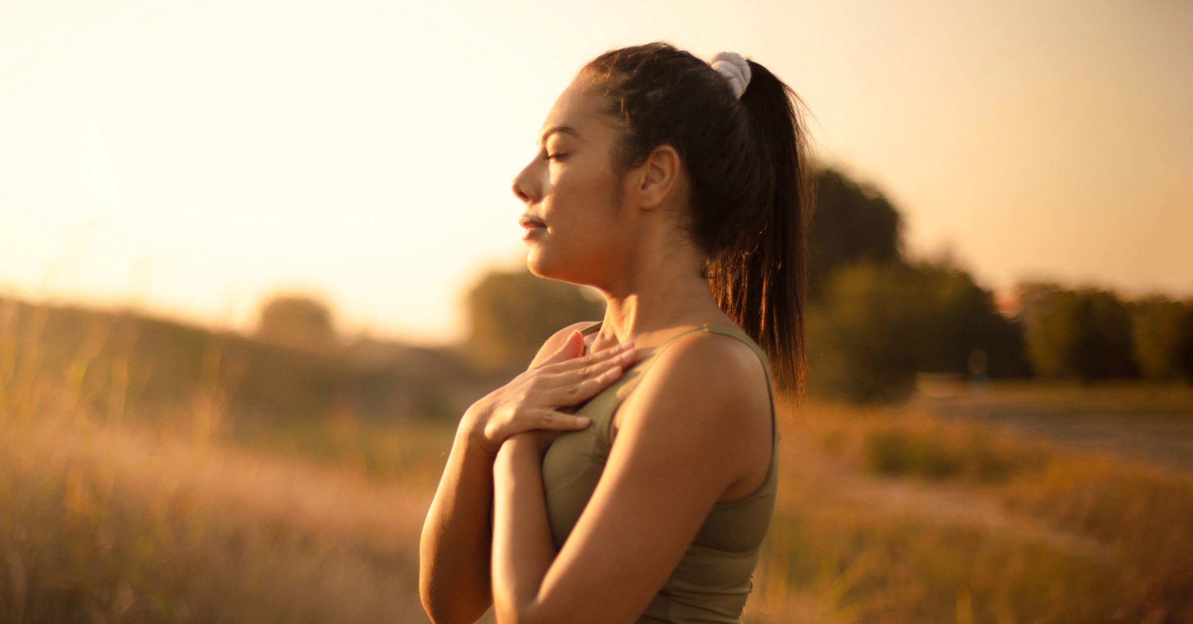 En kvinne står alene og nyter den friske luften ute på en eng ved soloppgangen, mens hun har øynene lukket og holder hendene på brystet.