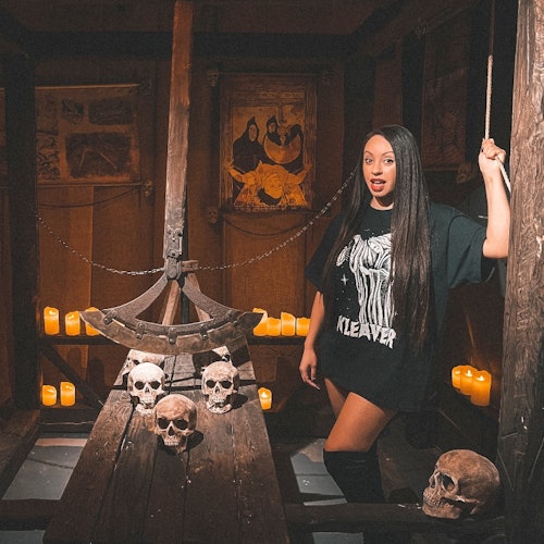 ਇੱਕ ਔਰਤ ਇਕ ਮਿੰਗ ਬੁੱਕ ਨਾਲ ਸਜਾਇਆ ਹੋਇਆ ਕਮਰਾ ਵਿੱਚ ਖੜੀ ਹੈ, ਜਿਸ ਵਿੱਚ ਮੋਮਬੱਤੀਆਂ, ਸਿਸਟੇਮ ਅਤੇ ਮੱਧਕਾਲੀਆ ਸਜਾਵਟ ਹੈ, ਇਕ ਰਸੇ ਨੂੰ ਲੱਕੜ ਦੇ ਡਿਵਾਈਸ ਦੇ ਨਾਲ ਫੜੀ ਹੈ।