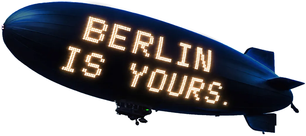 A dark blue blimp with glowing text on it reading "Berlin is yours" hovering in the air.