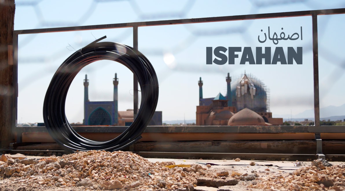 A coil of black plastic pipe resting on a rooftop, framing the domes and minarets of Isfahan's Imam Mosque — partly wrapped in scaffolding — seen through a hexagonal fence.