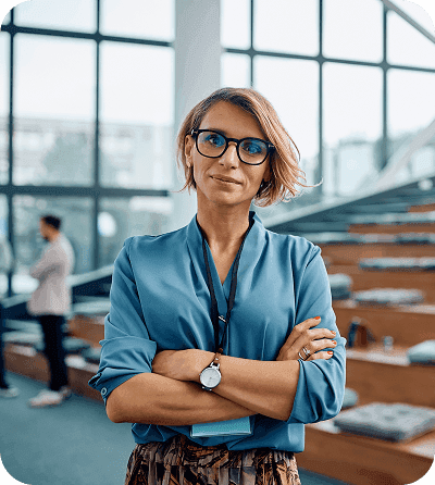“Woman with short wavy hair and glasses, wearing a blue blouse and a lanyard, standing with arms crossed in a modern office with large windows.”
