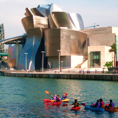 People kayaking in a river near a modern, metallic building with a unique architectural design under a clear sky.