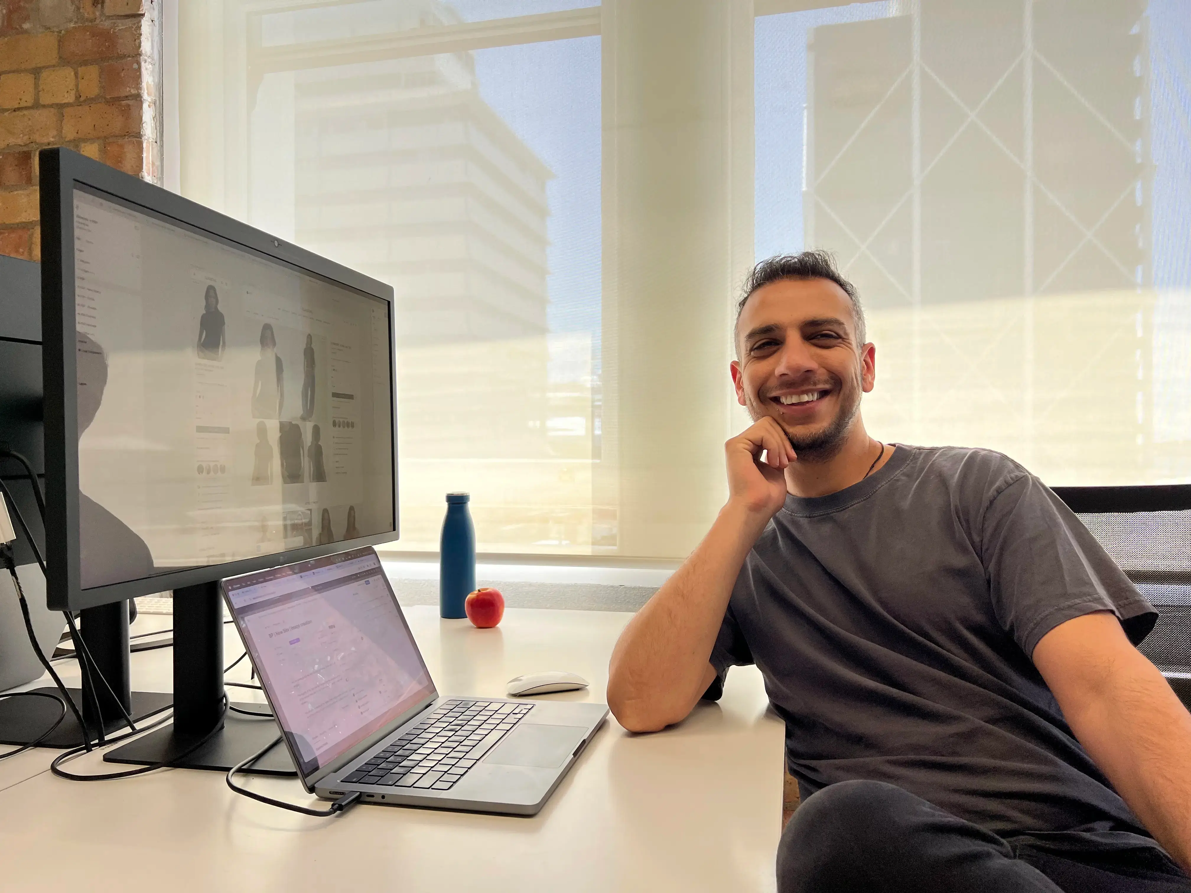 A smiling man with curly hair is sitting at a desk, looking at the camera, with a laptop and a lamp in the background.