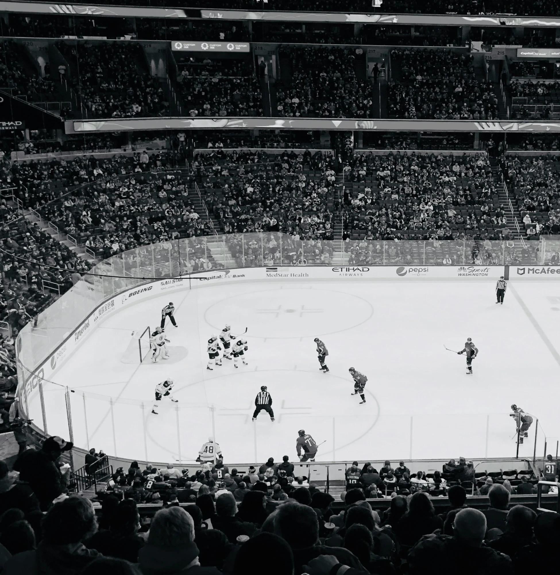 Black and white hockey game in progress, showing players on ice with packed arena stands in background. Professional sports atmosphere elevating the game experience.