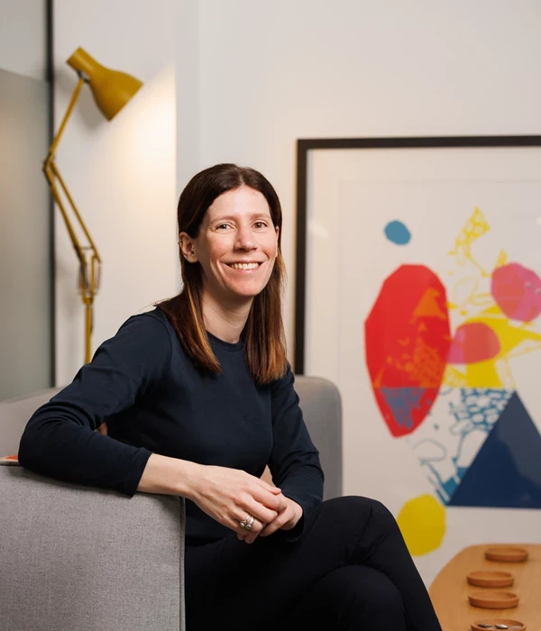A person seated on a light grey upholstered chair in a modern, softly lit indoor space. They are wearing a dark long‑sleeved top and dark trousers, sitting at an angle with their hands loosely clasped. Their straight, shoulder‑length brown hair is worn down. Behind them is a framed abstract artwork with bold red, yellow, blue, and black shapes, alongside a yellow adjustable floor lamp. A small round side table with wooden objects sits nearby, giving the setting a calm, contemporary studio or office feel.