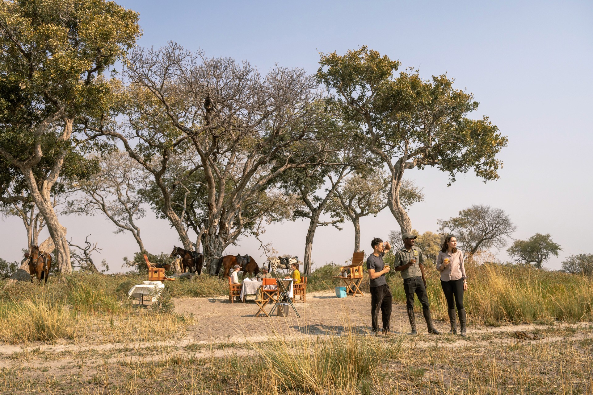 Kujwana Delta Ride, Okavango Delta, Botswana. Tre personer på ridsemester med uppdukat bord och hästar i bakgrunden.