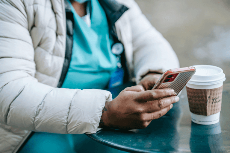 Care giver in scrubs using a smartphone while sitting at a table with a takeaway coffee.