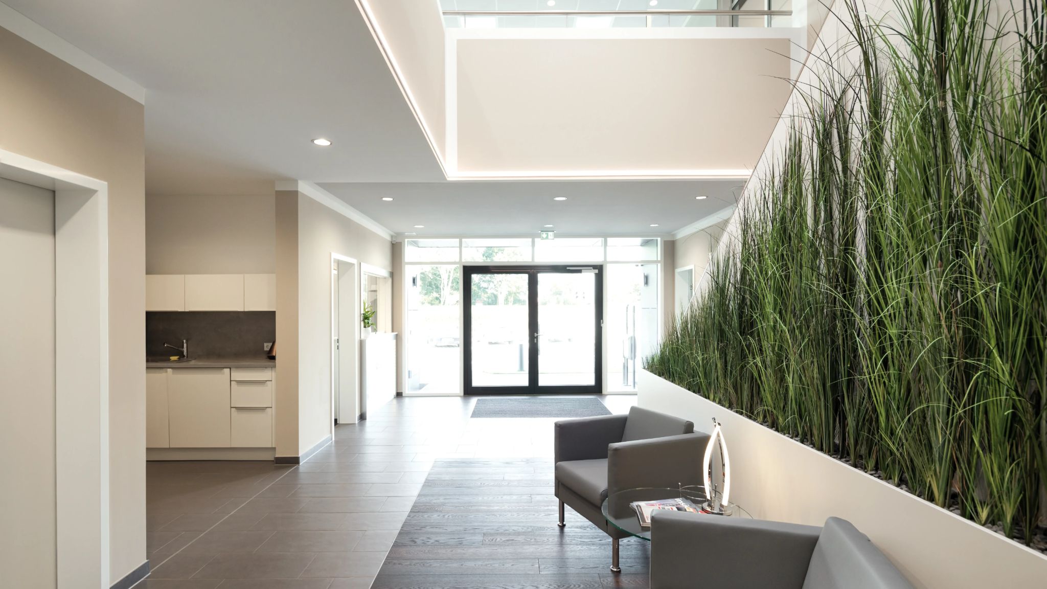 Modernes Foyer mit Blick auf Gläserne Eingangstür, Zimmerpflanzen, zwei graue Sessel und eine Büro-Küchenzeile.