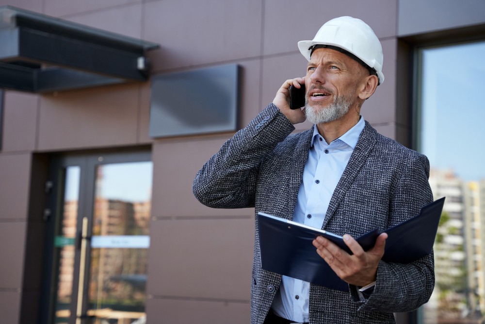 Contractor in a hard hat speaking on the phone outside a building, showing managed communications and contractor oversight.
