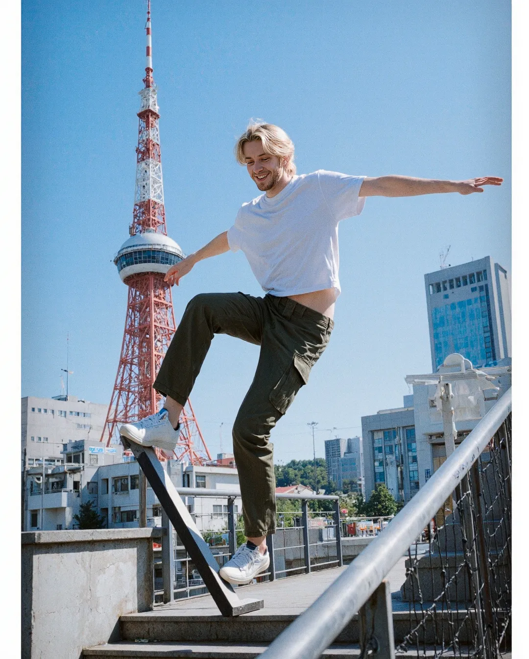 Person balancing on railing with Tokyo Tower in background, wearing white t-shirt and cargo pants against clear blue sky