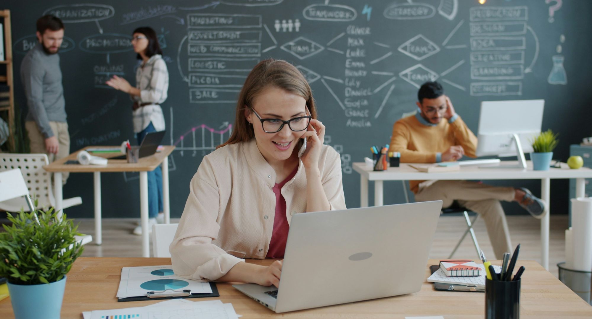 mulher sentada à mesa com notebooks e em ligação no celular numa sala de escritório com outras pessoas ao fundo