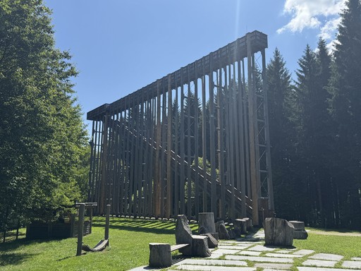 Tall, wooden structure surrounded by green trees and grass under a cloudy sky.