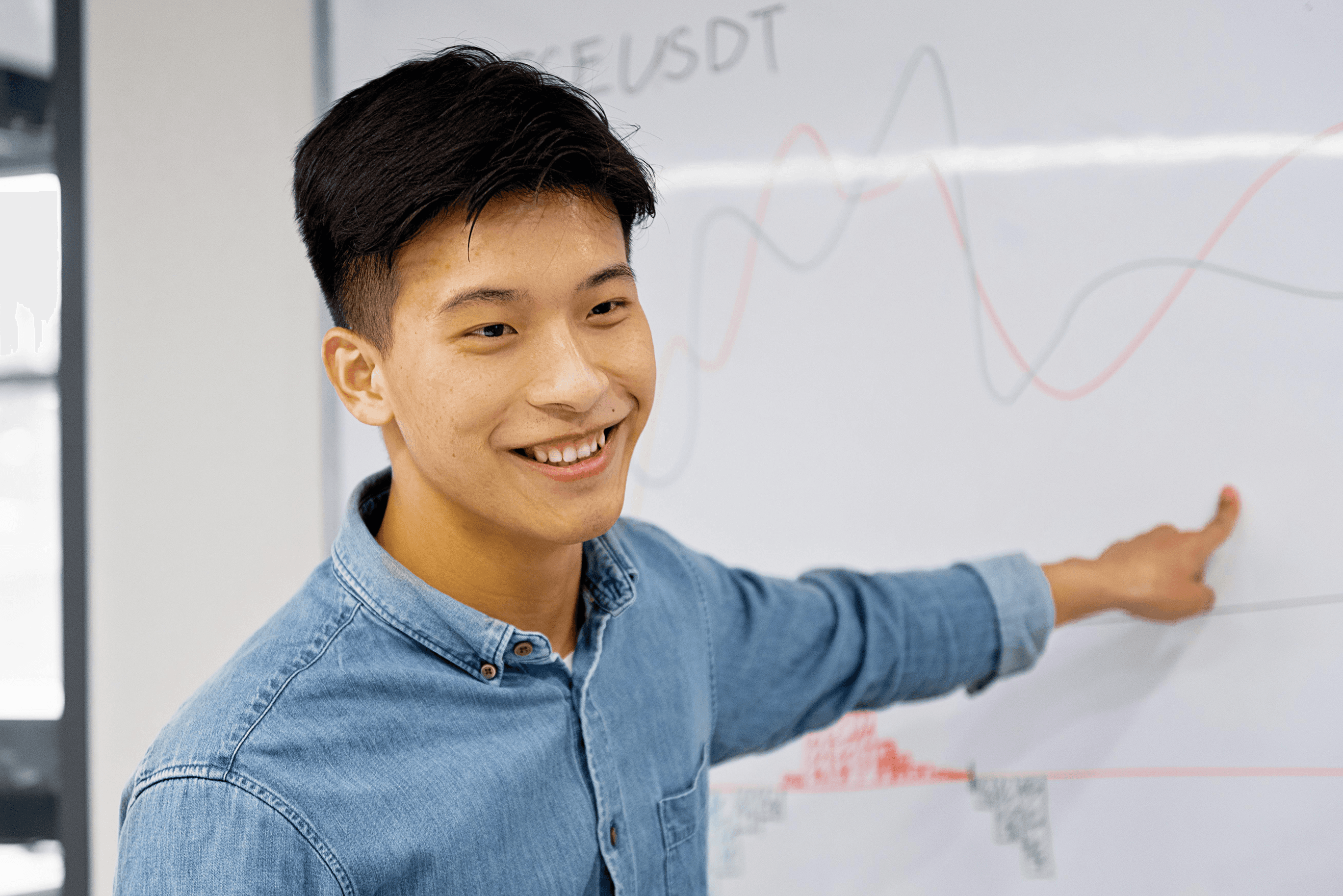 A person in a denim shirt is smiling while pointing at a financial chart on a whiteboard, featuring fluctuating graph lines and numerical data.