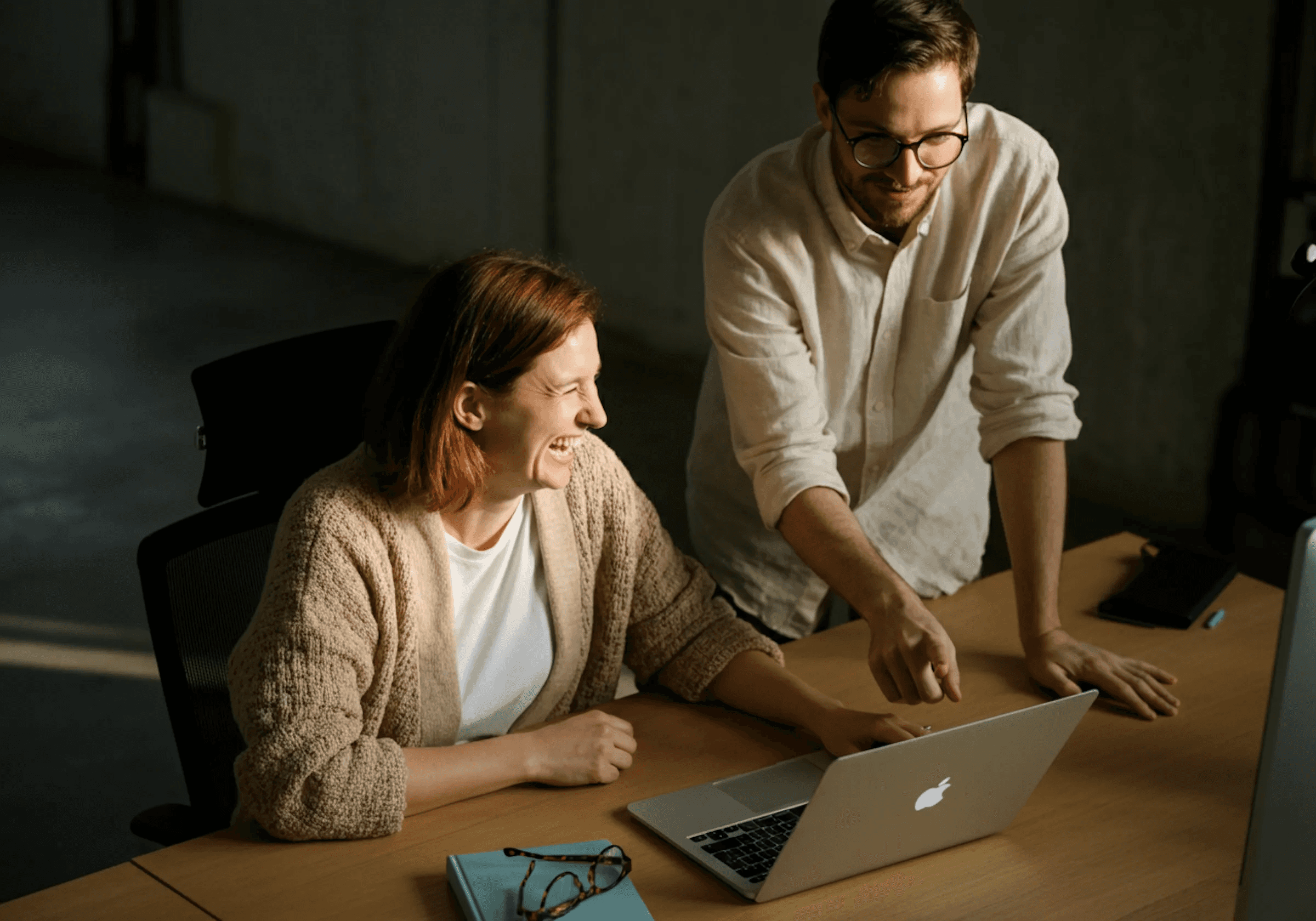 Woman smiling while seated at a desk with a laptop as a man in glasses leans over to show her something on the screen.