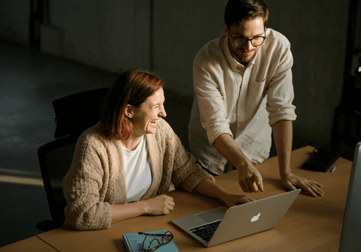 Woman smiling while seated at a desk with a laptop as a man in glasses leans over to show her something on the screen.