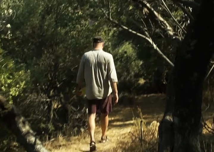 Άνδρας περπατά σε μονοπάτι μέσα σε ελαιώνα το καλοκαίρι, φορώντας καπέλο και κοντό παντελόνι.