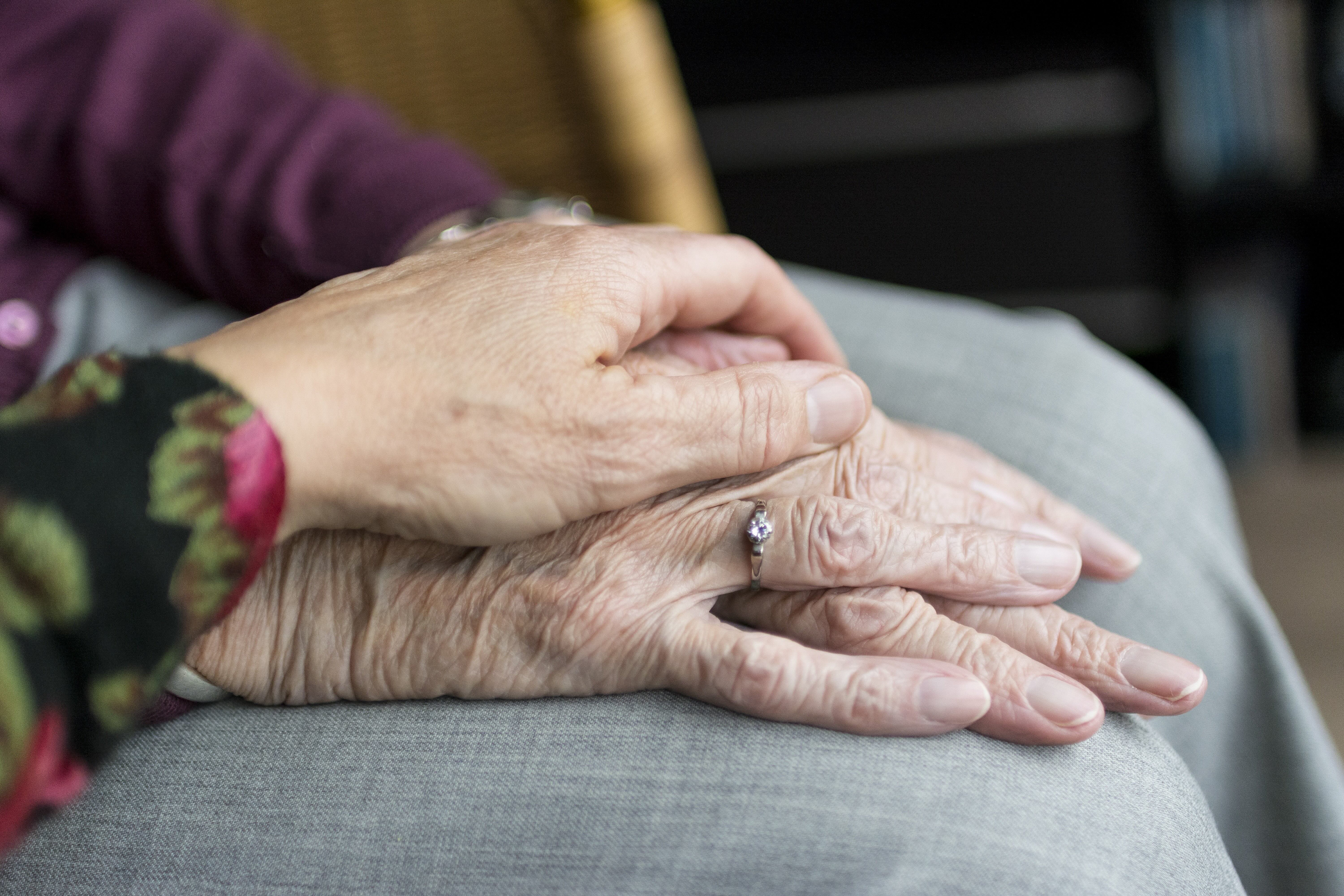 Two hands, one elderly and one younger, are gently clasped together, symbolizing support and connection; the elder's hand displays a diamond ring while resting on a gray-clad lap.