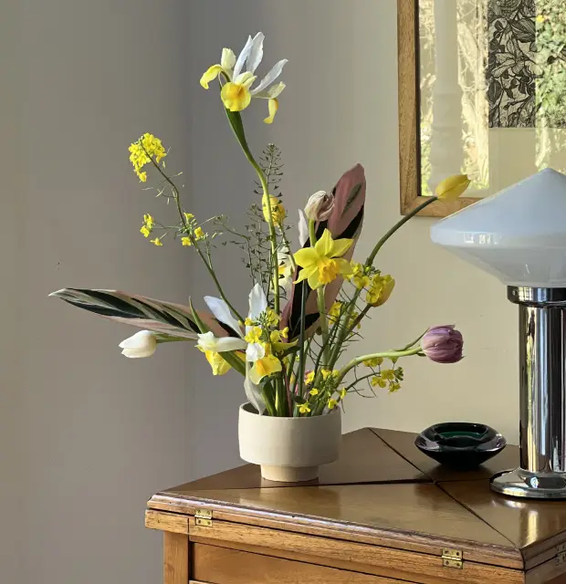 Sala luminosa con sillón blanco, cojines verdes y un aparador de madera decorado con un florero de ramas con flores rosas, cuadros en la pared y piso de madera natural