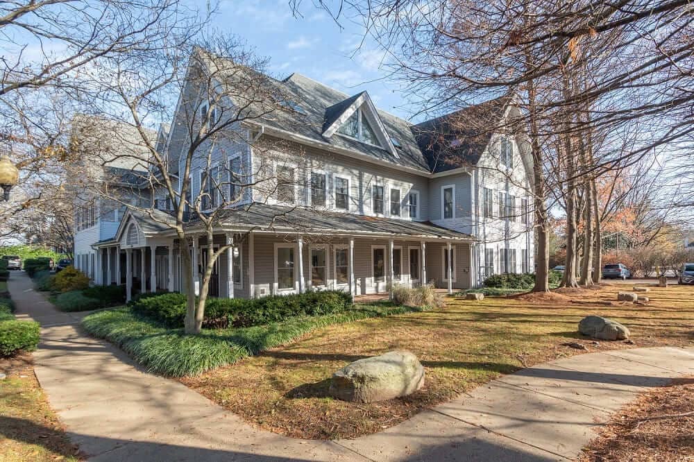 A large, multi-story house with gray siding and a porch, surrounded by trees and landscaped yard.