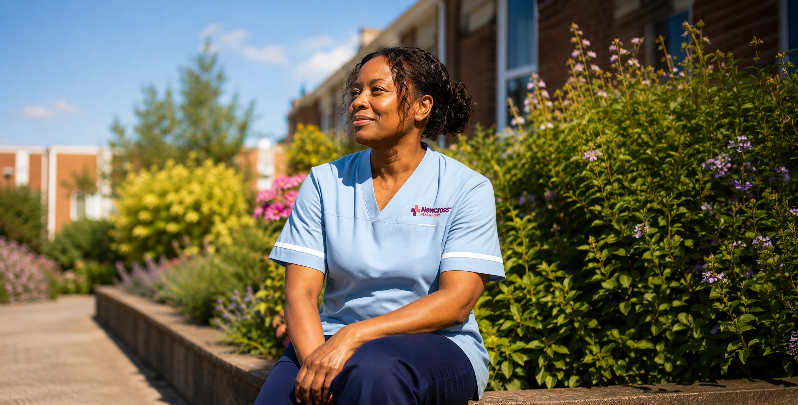 A Newcross Healthcare support worker in uniform sitting outside in a sunny garden, smiling and looking away from the camera