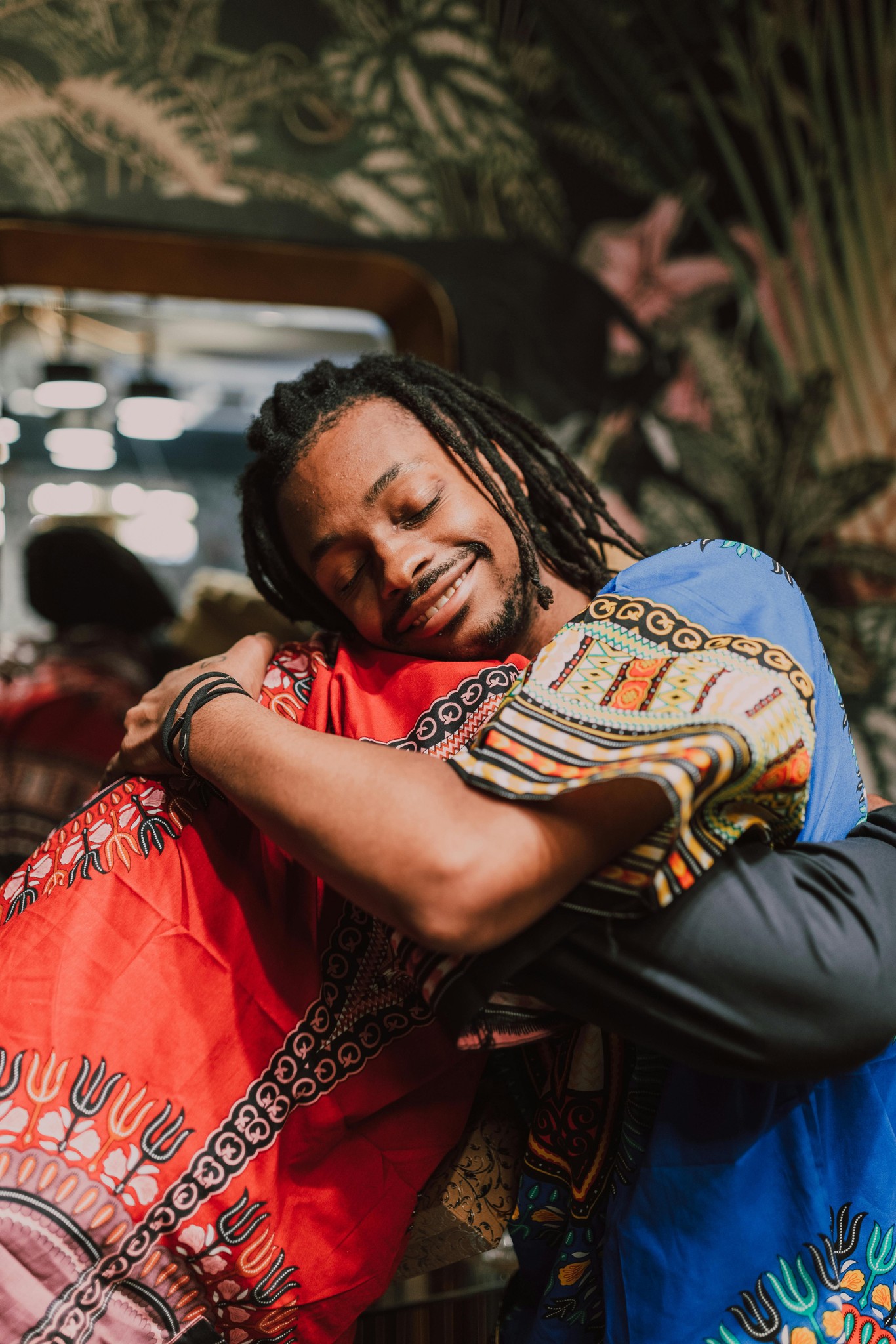 A warm embrace between two individuals wearing vibrant Dashiki garments. The focus is on a smiling young Black man with dreadlocks and closed eyes, leaning his head affectionately against the other person. He wears a blue Dashiki with intricate traditional sleeve patterns, wrapping his arms around his companion, who wears a striking red Dashiki with similar ornate detailing. The indoor background is softly lit with green patterned wallpaper.