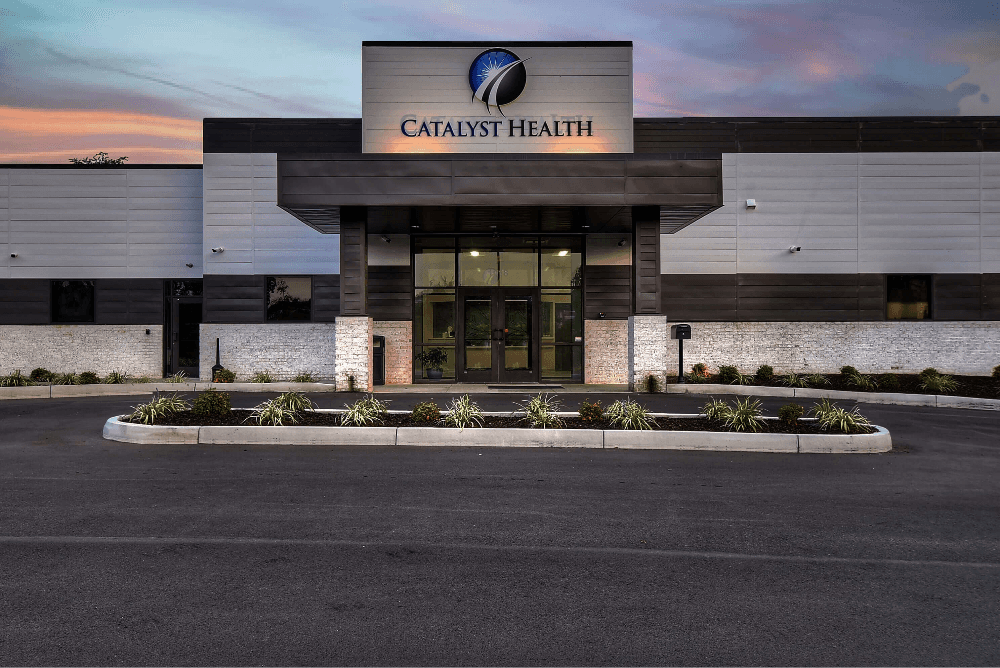 A modern building with a sign, surrounded by a paved area and some landscaping, under a colorful sky at dusk.