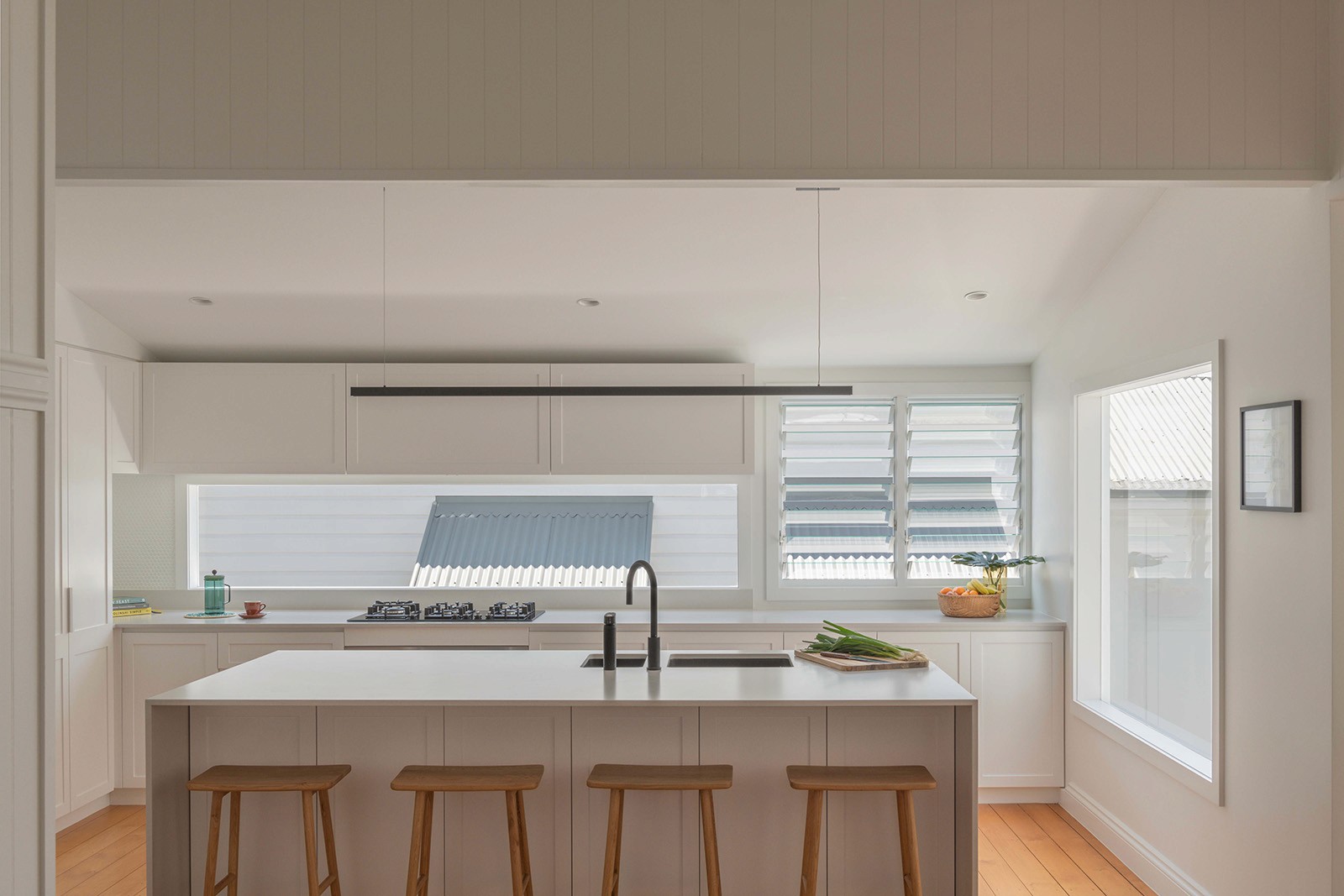 Minimal white kitchen with a long island, timber stools, skylight-style windows, and soft natural light throughout.