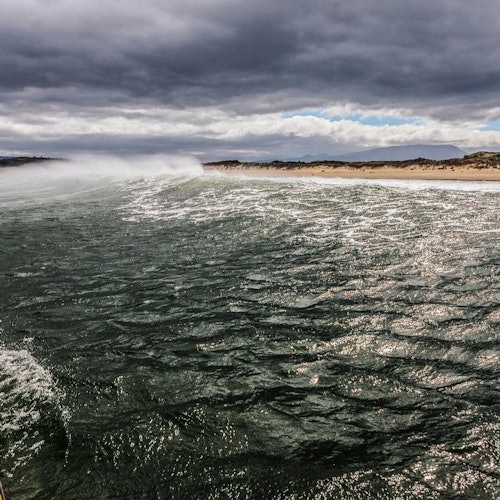 Тъмна, вълниста океанска вода под облачно небе с далечни пясъчни дюни по брега.