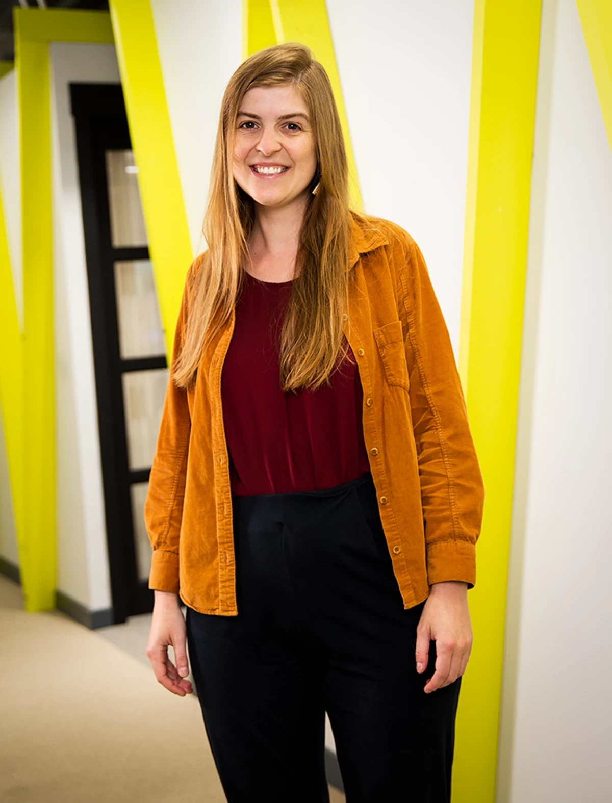 Person standing indoors against a wall with bright yellow diagonal accents, wearing a burnt orange button-up shirt over a dark red top and black trousers.