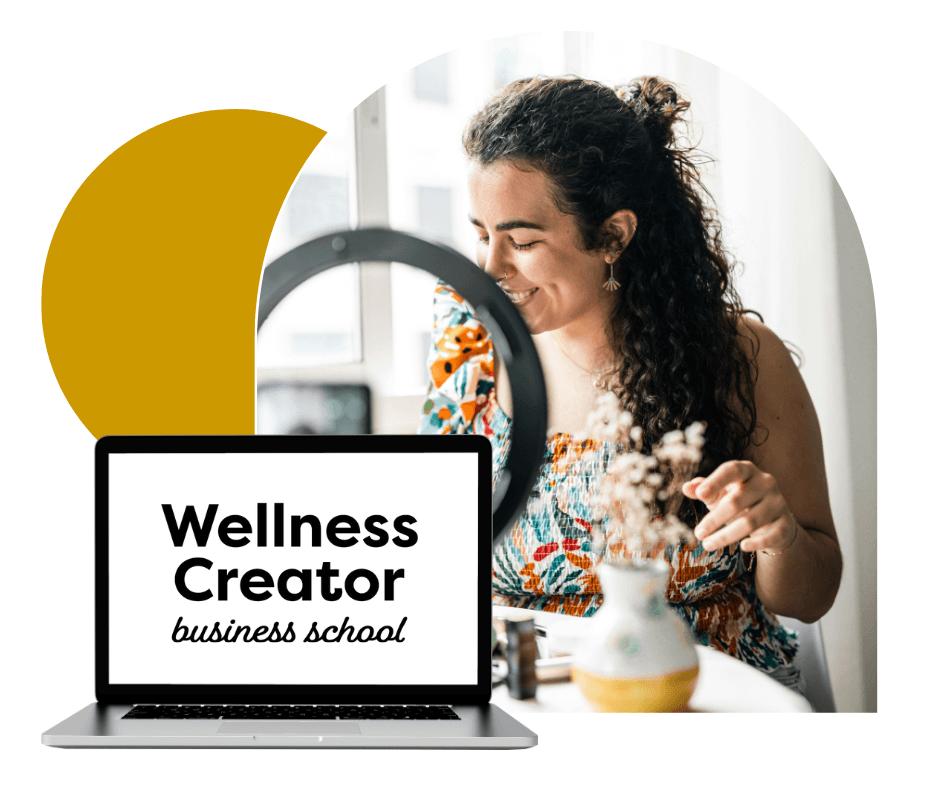 A person with curly hair smiles while seated at a desk next to a laptop displaying the text "Wellness Creator business school," with a ring light and decorative vase in the background.