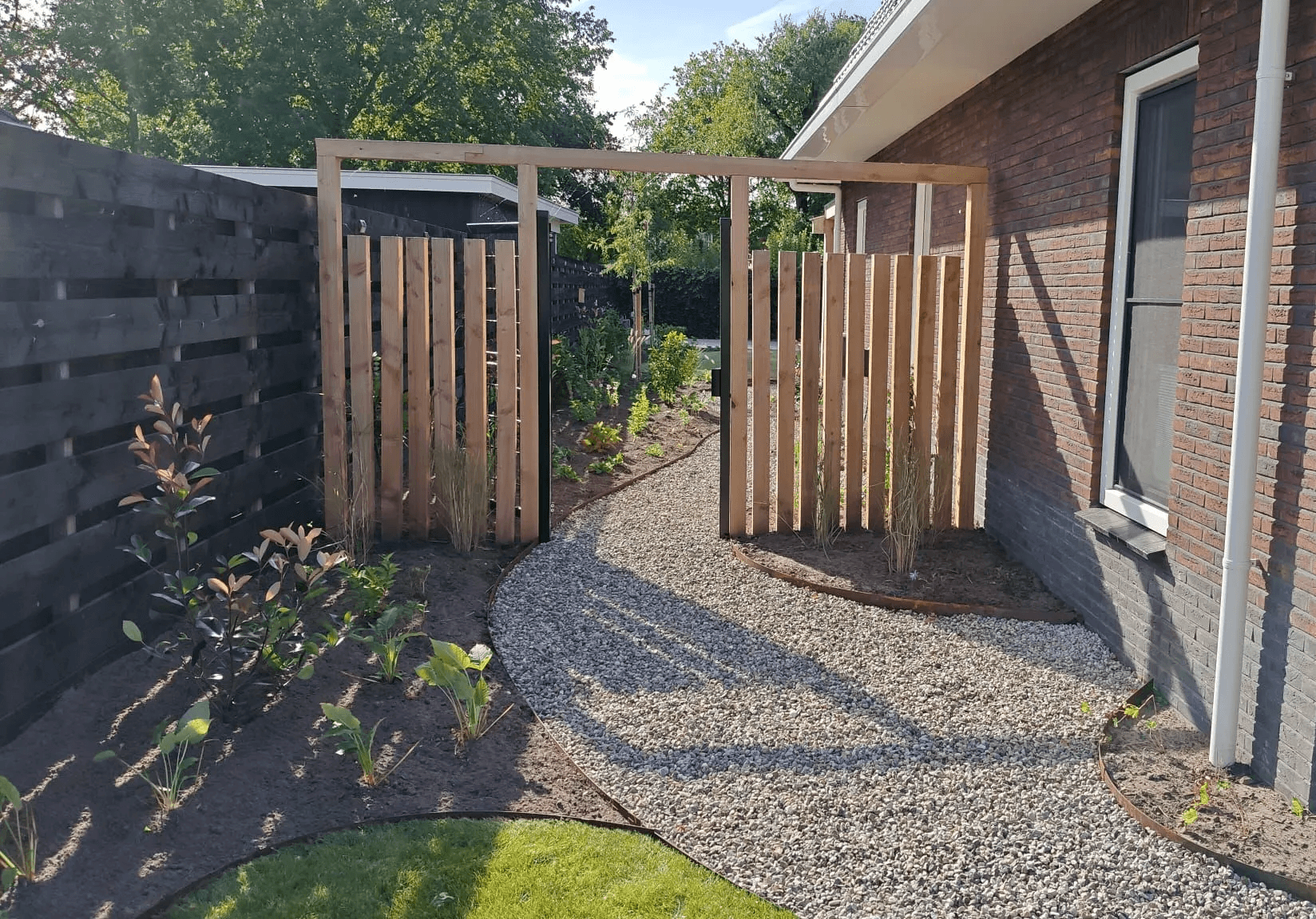 Smal tuinpad van grind met houten poort, jonge beplanting en zwarte schutting naast nieuwbouwwoning in Friesland.