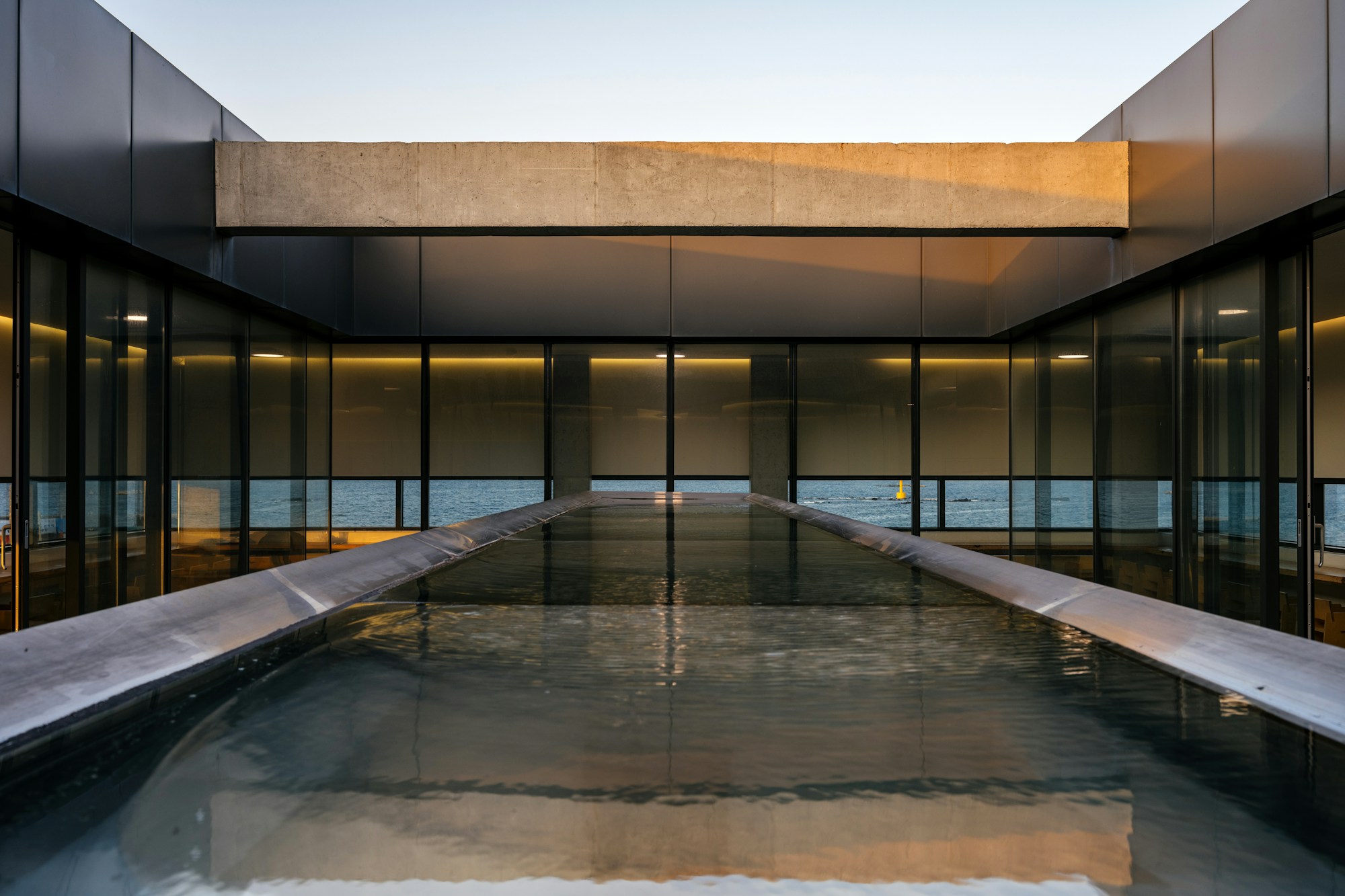 Glazed pavilion around a long reflecting pool, its roof slab lit by low evening sun