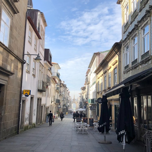 一條狹窄的鵝卵石街道，兩旁是老舊的建築，戶外咖啡桌上擺著陽傘，幾個人在藍天之下漫步。