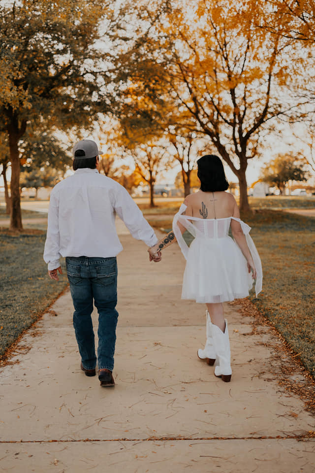 A couple in wedding attire walks hand in hand across a sunlit field.