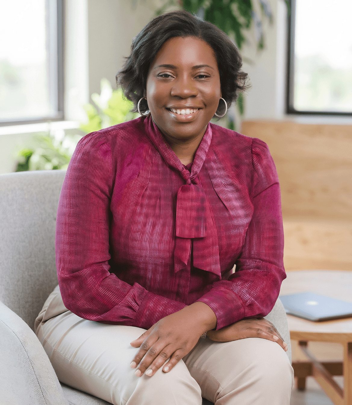 A woman in a burgundy blouse and beige pants sits on a gray chair, smiling warmly. A laptop is visible in the background on a wooden surface.