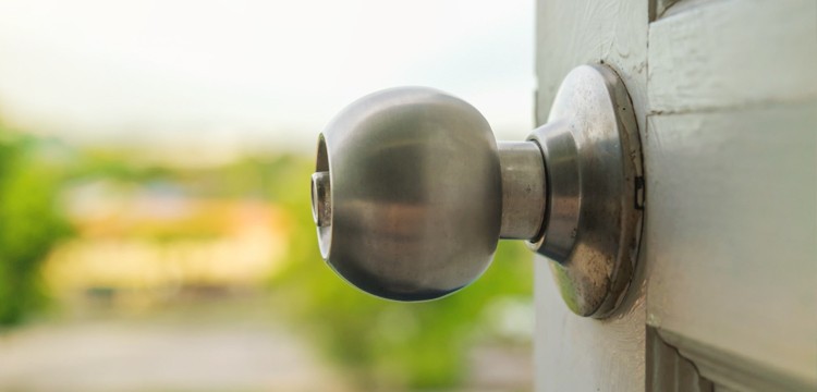 Side view close-up of a doorknob on a white wooden door, with a vivid green, sunny backdrop.