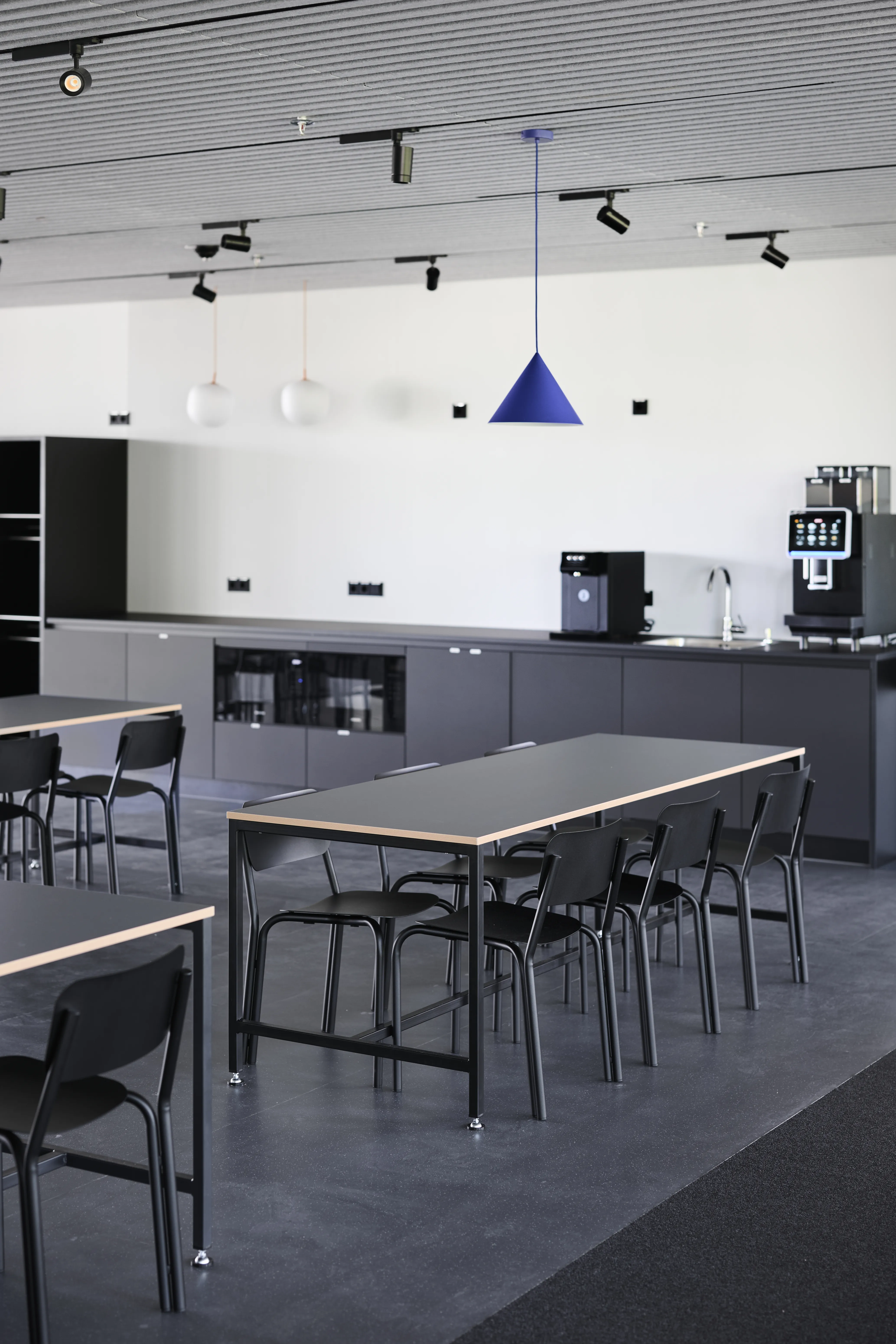 Dark kitchen counter at ENPAL with black cabinets, coffee machines, a cobalt cone pendant light, white globe lamps and black tables with matching chairs on dark flooring.