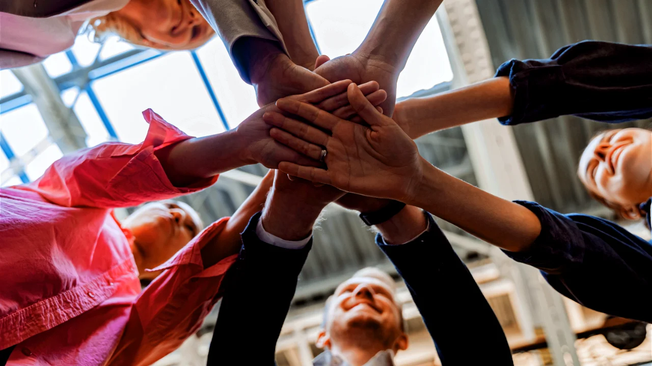 Un groupe de personnes souriantes, vues d'en bas, empilant leurs mains au centre en signe d'unité et de collaboration.