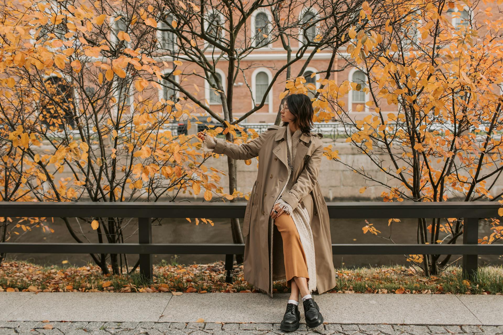 A woman in a trench coat sits on a bench surrounded by autumn leaves
