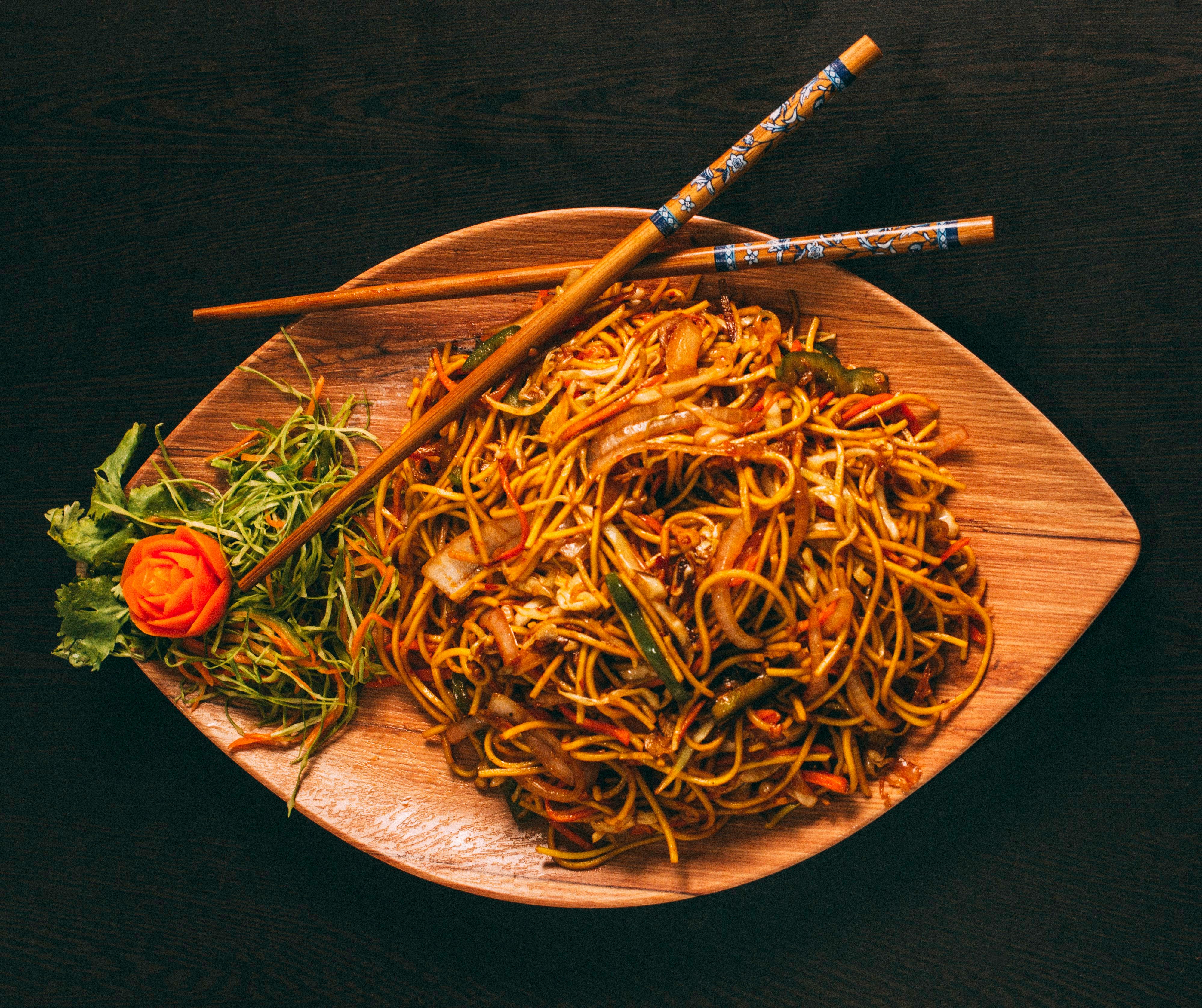 Stir-fried noodles with vegetables on a wooden platter with chopsticks and carrot garnish